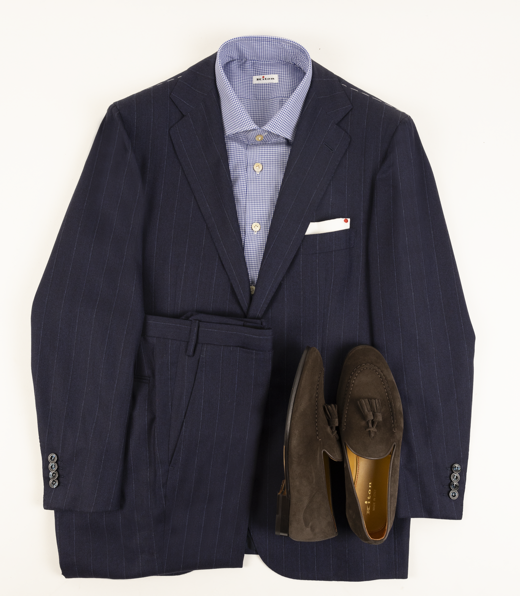 Kiton navy pinstripe suit laid flat with a blue checked shirt, white pocket square and brown suede tassel loafers